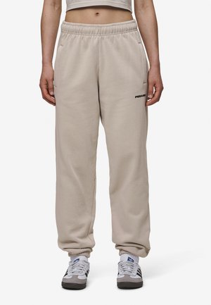 Person trägt beige Jogginghose mit elastischen Bündchen und Seitentaschen, kombiniert mit weißen und schwarzen Turnschuhen.