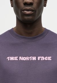 Nahaufnahme einer Person, die ein lila Rundhalsshirt mit pinkfarbenem, stilisiertem Schriftzug "THE NORTH FACE" auf der Brust trägt.
