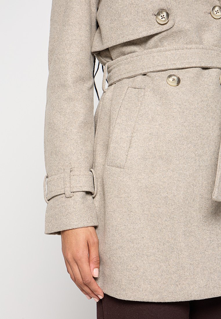 Trench-coat en mélange de laine de couleur beige clair avec de grands boutons, une taille ceinturée et de larges manches à revers. Il dispose de poches avant et d'une texture travaillée.