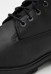 Botte en cuir noir avec bout arrondi, semelle en caoutchouc épaisse, œillets en métal et lacets noirs, avec des détails de couture visibles le long des côtés.