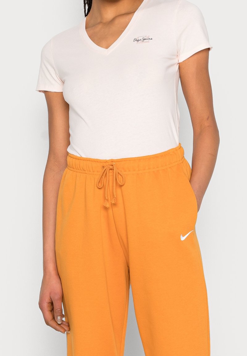 T-shirt rose clair à col en V associé à un pantalon de survêtement orange à taille élastique, arborant un logo Nike blanc sur la jambe gauche. Tissu doux et design décontracté.