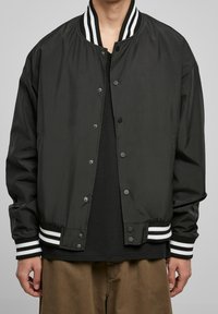 Czarna kurtka typu bomber wykonana z lekkiego materiału, z prążkowanym kołnierzem z białymi paskami, zapięciem na zatrzaski i elastycznymi mankietami.