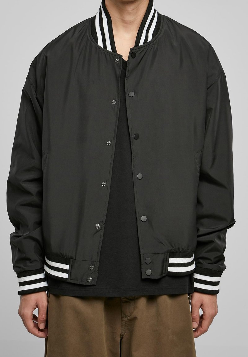 Czarna kurtka typu bomber wykonana z lekkiego materiału, z prążkowanym kołnierzem z białymi paskami, zapięciem na zatrzaski i elastycznymi mankietami.
