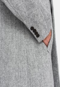 Abrigo texturizado en gris de espiga con un solo bolsillo y cuatro botones oscuros en el puño, que muestra un diseño a medida.