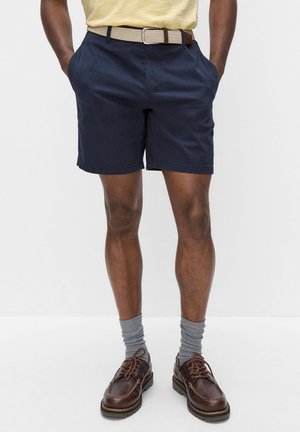 Man draagt marineblauwe shorts, beige gevlochten riem, grijze sokken en bruine leren veterschoenen, staat met handen in de zakken.