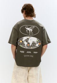 T-shirt oversize verde oliva con stampe grafiche bianche di una mosca e una mappa del mondo, con testo sottostante. Maniche corte e vestibilità rilassata.