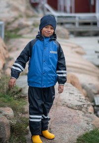 Garçon portant une combinaison de pluie bleue et noire avec des bandes réfléchissantes, un chapeau marine et des bottes de pluie jaunes, debout sur un chemin rocheux à l'extérieur.