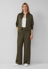 Olivgrüne Satin-Bomberjacke mit hohem Kragen, kombiniert mit passenden weiten Hosen. Ein weißes Top und weiße Sneaker runden den Look ab.