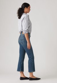 Donna con i capelli ricci raccolti in un ponytail, che indossa una camicia a righe, jeans blu a vita alta e ballerine nere a punta, in posa di profilo su uno sfondo neutro.