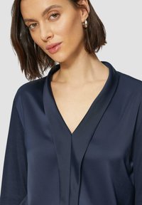 Marine satijnen blouse met een diepe V-hals en losse mouwen. De stof heeft een zachte textuur die het minimalistische ontwerp benadrukt.