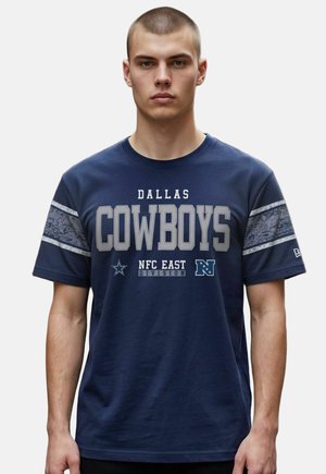Młody mężczyzna z krótkimi włosami, noszący granatowy T-shirt Dallas Cowboys z dywizji NFC East z szarymi paskami na rękawach.