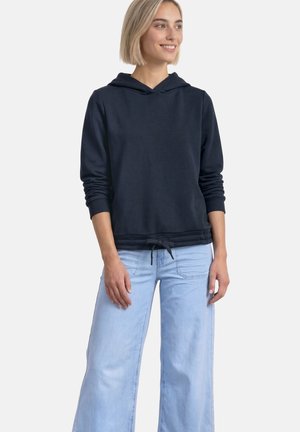 Femme portant un sweat à capuche bleu marine avec cordon de serrage à l'ourlet et un jean large bleu clair, debout et souriante, légèrement tournée vers la droite.