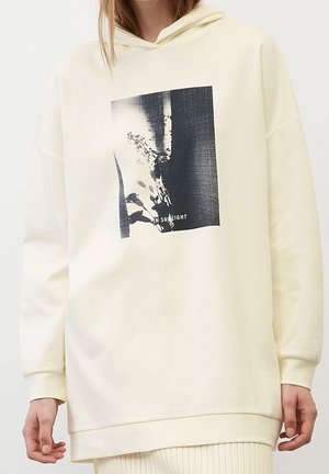 Personne portant un sweat à capuche écru avec un imprimé abstrait noir et blanc et les mots "IN SUNLIGHT" sur le devant.