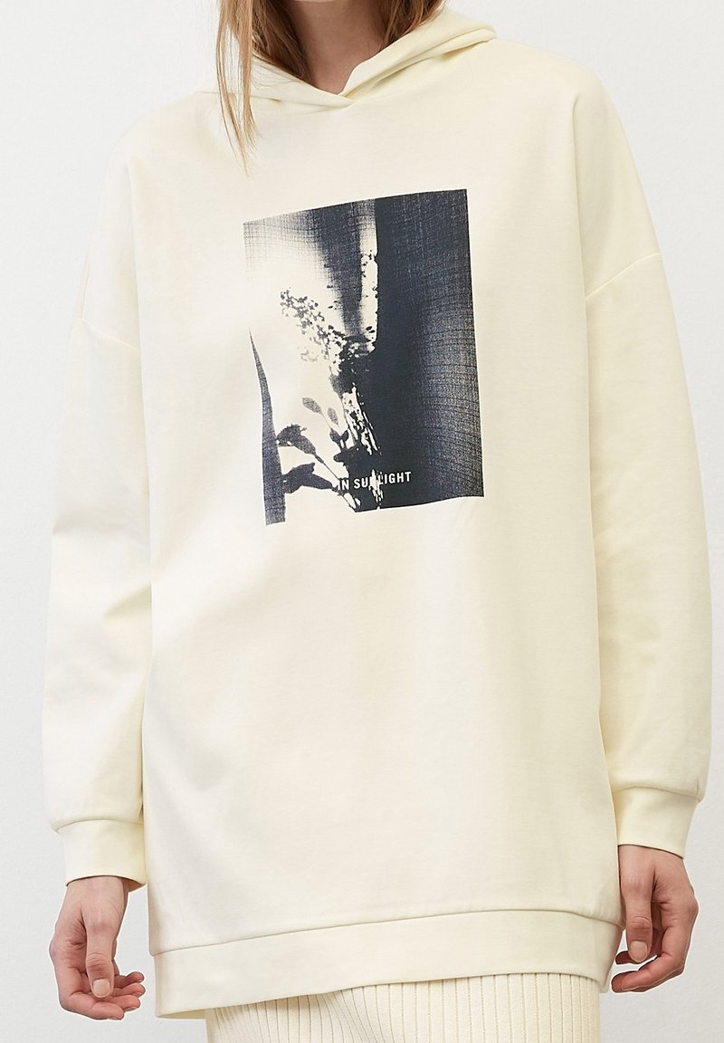 Personne portant un sweat à capuche écru avec un imprimé abstrait noir et blanc et les mots "IN SUNLIGHT" sur le devant.