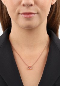 Roségold-Halskette mit einem runden Anhänger, der einen klaren Stein zeigt, auf einer dünnen Kette, präsentiert gegen eine dunkle Bluse mit Kragen.