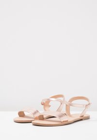 Sandali metallici oro rosa con due ampie cinghie sui piedi e una sottile cinghia alla caviglia, caratterizzati da una texture in pelle liscia e suola piatta.