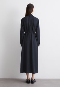 Abito a maniche lunghe, lungo fino al pavimento, in tessuto blu scuro con vita aderente. Presenta una texture liscia e un classico colletto da camicia. Indossato con scarpe nere.