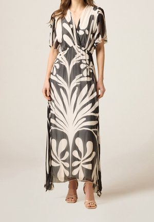 Femme portant une robe maxi noire et blanche avec des motifs floraux abstraits, des manches courtes flottantes et des escarpins nus à bout ouvert, debout sur un fond uni.