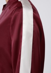 Μπουφάν από σατέν μπορντό με λευκή ρίγα από σατέν κατά μήκος του ώμου. Το ύφασμα έχει ομαλή υφή με ελαφριά λάμψη.