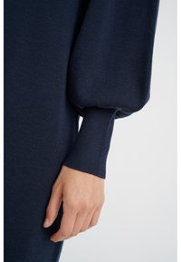 Maglione blu scuro con un tessuto a maglia texturizzata, maniche a sbuffo oversize e polsini aderenti. Ingrandimento di una mano appoggiata sul capo.
