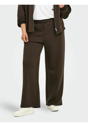 Personne portant un pantalon de survêtement marron foncé à jambes larges, des baskets blanches et une veste zippée assortie sur une chemise blanche, debout sur un fond uni.