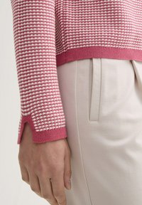 Strickpullover mit einem rosa-weißen Streifenmuster, gerippten Bündchen und einem leichten Schlitzdetail, kombiniert mit hellen, maßgeschneiderten Hosen.