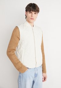 Vest crem cu matlasare, cu guler înalt și nasturi tip snap, purtat peste un pulover texturat maro și blugi de culoare albastru deschis.