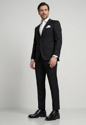 Mężczyzna stojący w czarnym garniturze, białej koszuli, czarnych eleganckich butach i srebrnym zegarku na nadgarstku na jasnym jednolitym tle.