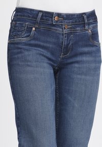 Jeans de mezclilla azul oscuro con un diseño clásico de cinco bolsillos, que cuentan con herrajes en tono cobre y un ligero desgaste cerca de los muslos.