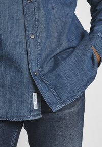 Camicia in denim blu scuro con design a bottoni, trama sottile e etichetta sul bordo inferiore. Indossata sopra jeans scuri.