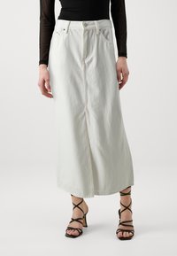 Jupe maxi en denim blanc avec une fente avant, fermeture à boutons et passants de ceinture ; portée avec des talons noirs à lanières. Texture lisse, design minimaliste.