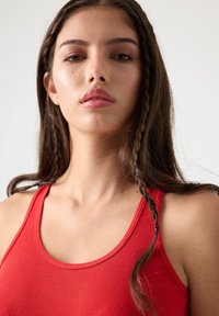 Top sin mangas de color rojo con cuello redondeado. La modelo tiene el cabello largo y ligeramente ondulado, con una sección trenzada, y un fondo neutro.