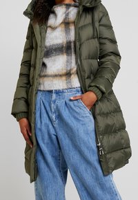 Olivenfarvet polstret jakke med hætte, med ribbede manchetter, lynlås og sidelommer, lagdelt over en grå og tanternet sweater og blå brede bukser.