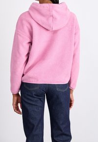 Persoon in een roze fleece hoodie en donkerblauwe jeans, van achteren gezien, staand tegen een plain witte achtergrond.