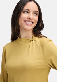 Długi rękaw w musztardowym kolorze, z kołnierzem typu mock oraz marszczeniem na ramieniu. Gładki, miękki materiał o dopasowanym kroju.