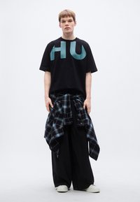 Μαύρο μπλουζάκι με μεγάλο ακουα χρωματιστό σχέδιο "HU", συνδυασμένο με άνετο, μαύρο παντελόνι. Μαύρο και μπλε καρό πουκάμισο δεμένο γύρω από τη μέση. Λευκά αθλητικά παπούτσια.