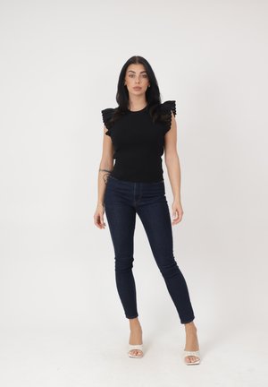 Kobieta stojąca, ubrana w czarny, falbaniasty, bezrękawnik, ciemne obcisłe dżinsy, białe sandały na obcasie z odkrytymi palcami oraz bransoletkę na lewej ręce.