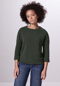 Grön, texturerad polotröja med rund halsringning och trekvartslånga ärmar, parat med denimjeans. Mjuk, avslappnad design.