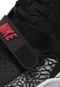 Baskets noires avec une tige en cuir texturé, un logo Nike rouge bien en vue, une sangle pour un maintien sécurisé et une section grise à motifs contrastants.
