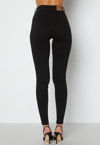 Černé úzké džínsy s vyšším pasem, hladkou strukturou, dvěma zadními kapsami a hnědou koženou nášivkou na pase. Kombinováno s černými páskovými podpatky.