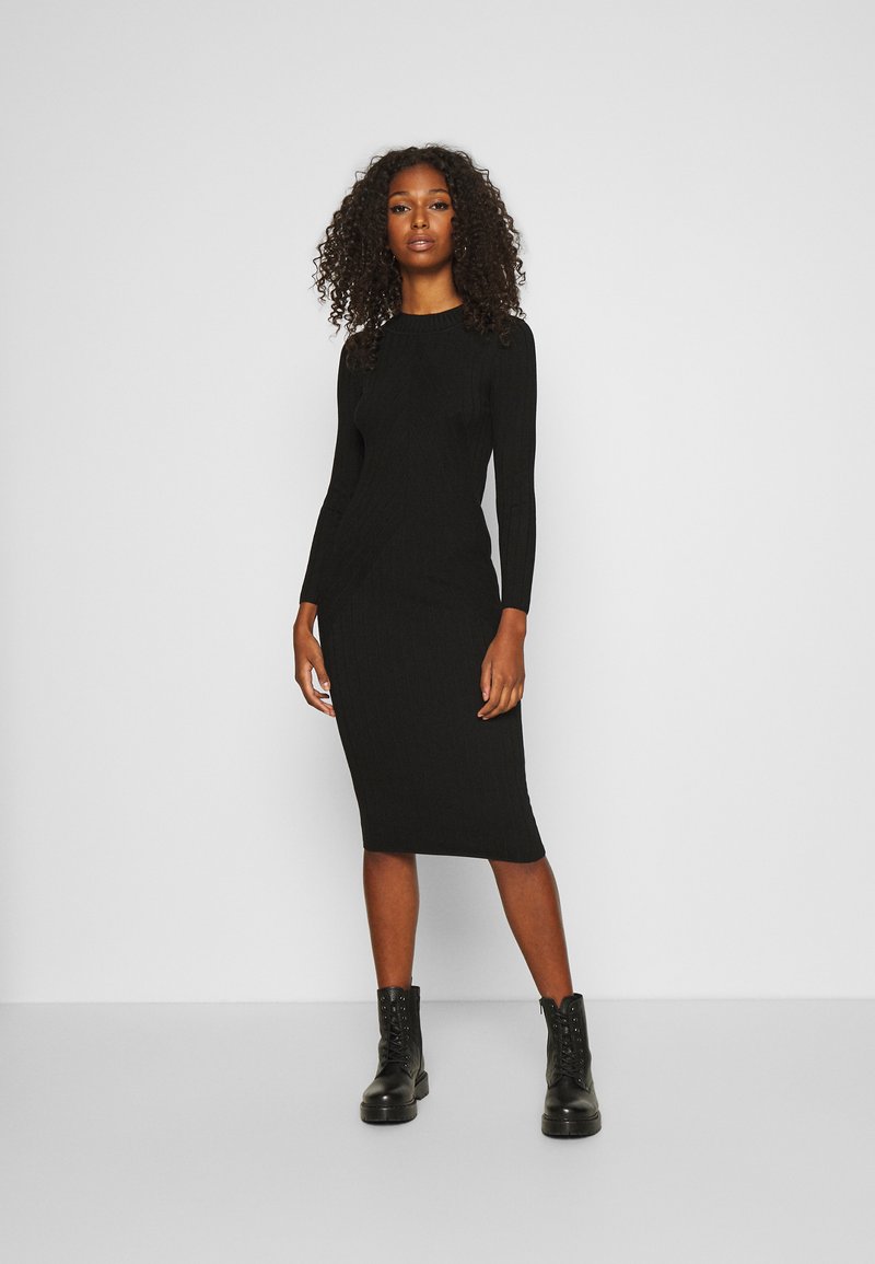 Črna rebrasta midi obleka z dolgimi rokavi, ki se ponaša z oprijetim silhueto in teksturiranim vzorcem. Upoštevana s črnimi čevlji za boj z vezalkami.