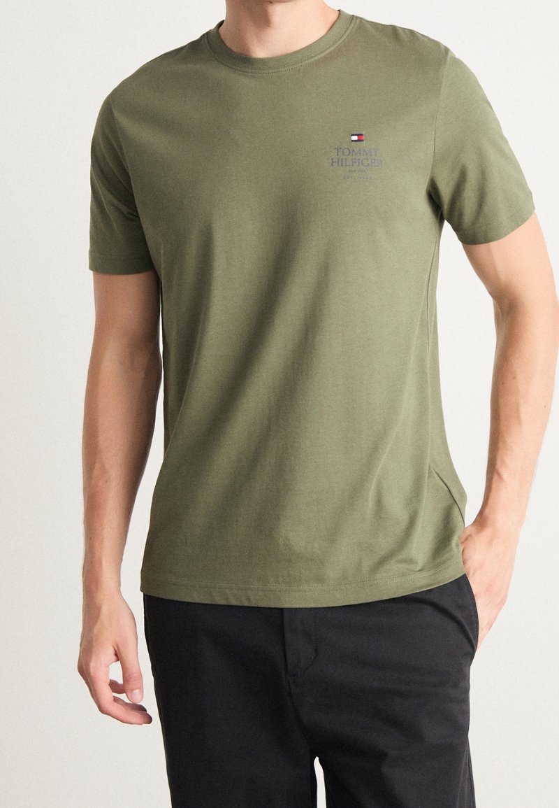 Camiseta de algodón verde oliva con cuello redondo, mangas cortas y un pequeño logo en el pecho. Presenta un diseño liso y una textura ligera.