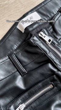 Κοντινό πλάνο μαύρων δερμάτινων παντελονιών με ασημένια φερμουάρ και θηλιά για ζώνη, τοποθετημένα σε ξύλινη επιφάνεια, με ετικέτα ρούχου στο εσωτερικό.