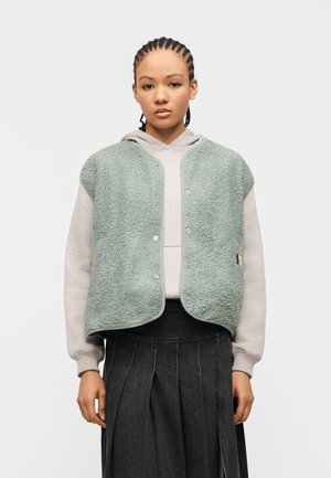 Junge Frau mit geflochtenem Haar, die einen hellgrauen Kapuzenpullover, eine minzgrüne Fleeceweste mit Druckknöpfen und einen dunklen, plissierten Rock vor weißem Hintergrund trägt.