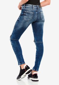 Schlanke Jeans in mittlerem blauen Denim mit distanziertem Design, fünf Taschen und Reißverschlussakzenten an den Seiten; kombiniert mit schwarzen Turnschuhen.