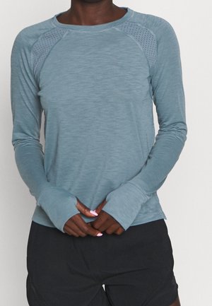 Persoon draagt een lichtblauw sportshirt met lange mouwen en duimgaten, en zwarte korte broek, staat met de handen gevouwen.