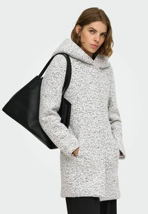 Jeune femme en manteau gris à capuche, les mains dans les poches, porte un grand sac cabas en cuir noir sur l'épaule.