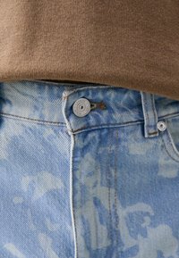 Jeans de mezclilla azul claro con un patrón desgastado y texturizado, que presentan un botón plateado y detalles de costura visibles. Tela marrón en la parte superior a la altura de la cintura.
