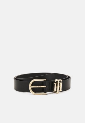 Ceinture en cuir noir avec une boucle en métal doré et des initiales décoratives dorées "TH" près de la boucle, enroulée sur fond blanc.