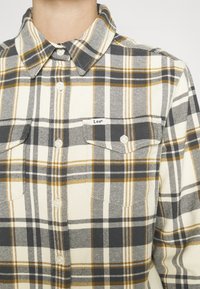 Homme portant une chemise à carreaux jaune, noire et blanche boutonnée, avec un col pointu et deux poches poitrine, dont une avec une petite étiquette "Lee".